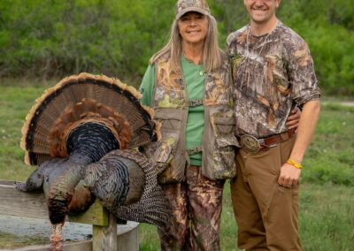 Two people, dressed in camouflage hunting gear, stand outdoors beside a large turkey they have hunted. The older woman on the left wears a camouflage hat and vest, while the young man on the right wears a camo shirt and brown pants. They're standing in a grassy area with trees in the background.