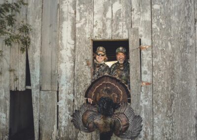 Two people in camouflage clothing look out from a weathered wooden structure, smiling. In front of them, a large turkey with its tail feathers fanned out is draped, suggesting a successful hunt. The wooden building has peeling paint and a rustic charm.