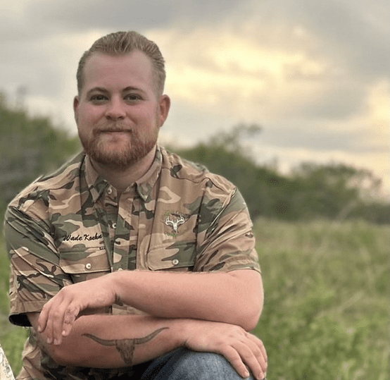 A man with short hair and a beard is wearing a camouflage-patterned shirt with an embroidered name, sitting outdoors in a field. His arms are crossed, and the background features greenery and a cloudy sky.