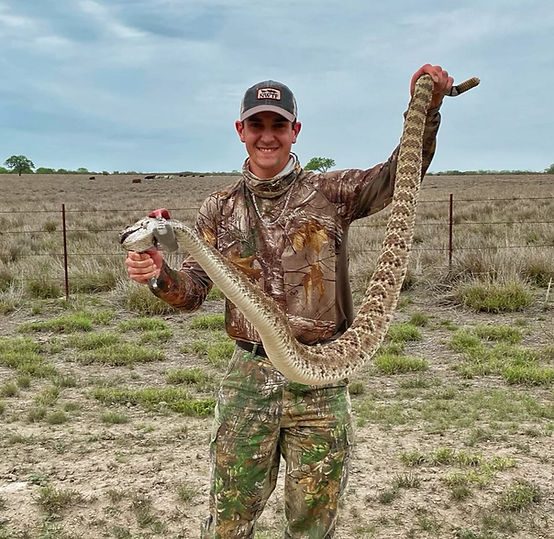 A person wearing camouflage clothing and a cap is standing in a dry, grassy area, proudly holding up a large rattlesnake by its tail. The background includes a wire fence and some vegetation.